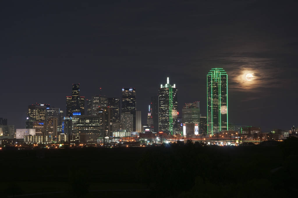 The Dallas county Texas skyline at night with a full moon from the Trinity River Greenbelt Park and picture provided by Prestige Car & Limo dallas texas.